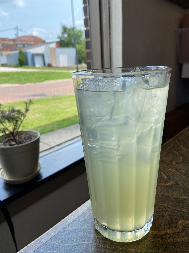 A tall clear glass with light yellow lemonade sits on a table. There is a window with a plant on the windowsill in the background. Photo by Jessica Hammie.