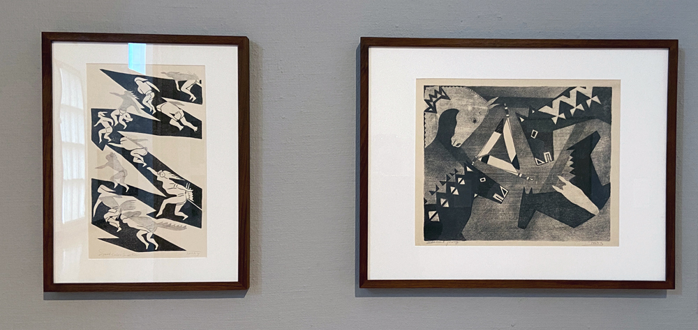 Julius Land Elk Twohy (Two-vy-nah-up), Speed, Color, Action on the left and Blessed Pony on the right, both lithographs ca. 1940. Two framed lithographs are mounted on a gray gallery wall. On the left, Speed, Color, Action features a black zig-zag with various figures. On the right, Blessed Pony features different horse heads in various states of profile circled around three knives set in a triangle toward the middle of the competition. Both lithographs are in black, white, and gray. Photo by Jessica Hammie. 