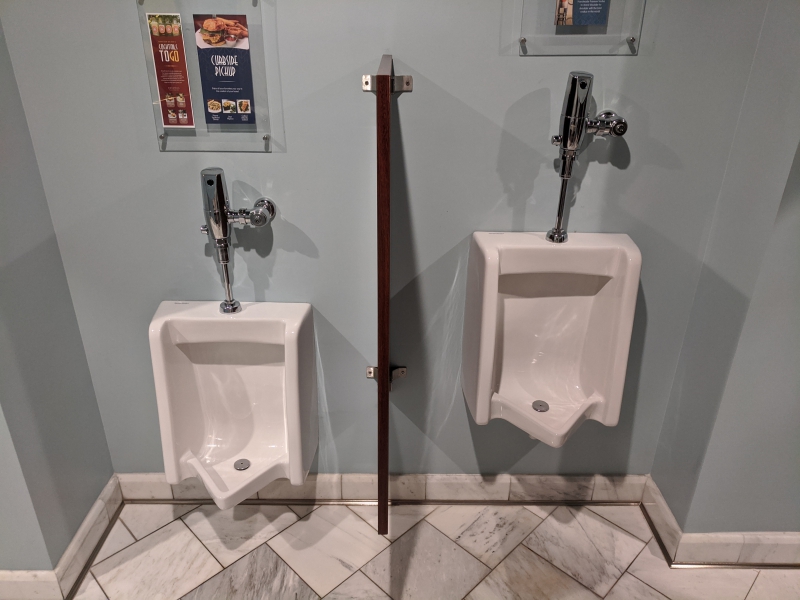 Two white urinals are side by side on a gray wall with a divider in between. The floor has square gray and white marble tile. Photo by Tom Ackerman.