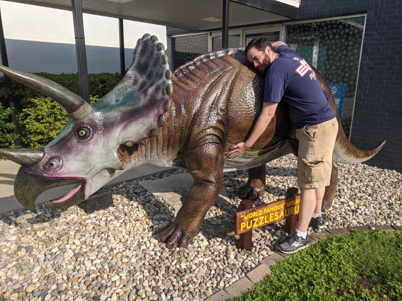 The author is wearing khaki shorts and a navy blue t-shirt. He is leaning into and hugging a large statue of a triceratops. Photo by Andrea Black.
