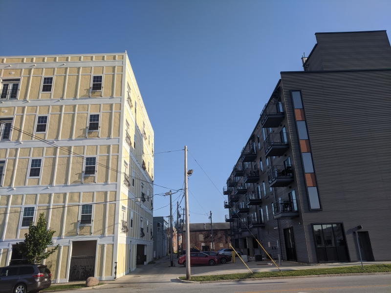 Two apartment buildings frame an alley. The building on the left is yellow with white trim and small rectangular windows. The building on the right is brown with balconies. Photo by Tom Ackerman.