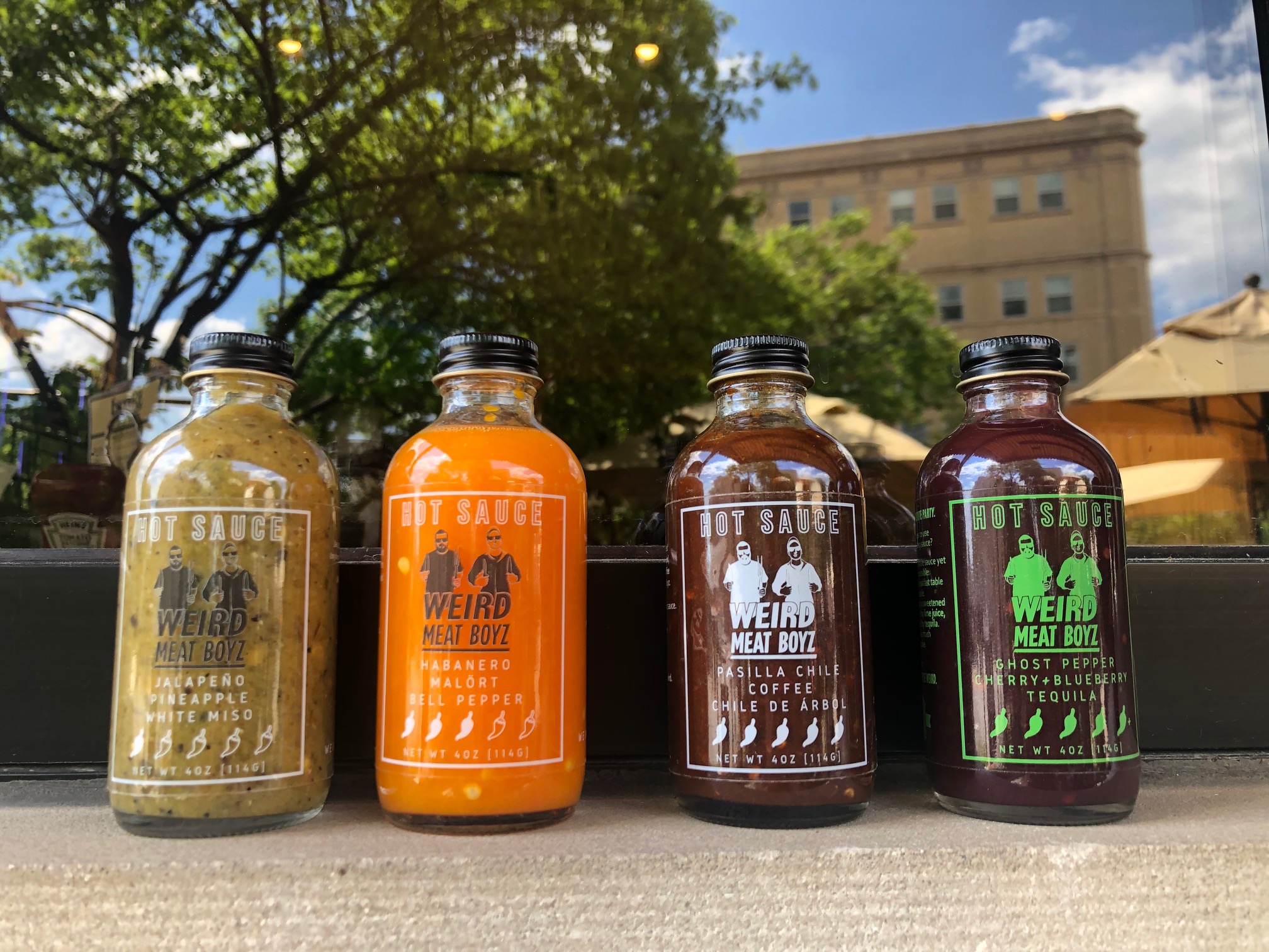 On a window ledge, there are four miniature bottles of hot sauce by Weird Meat Boyz. In the window behind, there are trees in Downtown Champaign and a bright blue sky reflected in the window. Photo by Alyssa Buckely. 