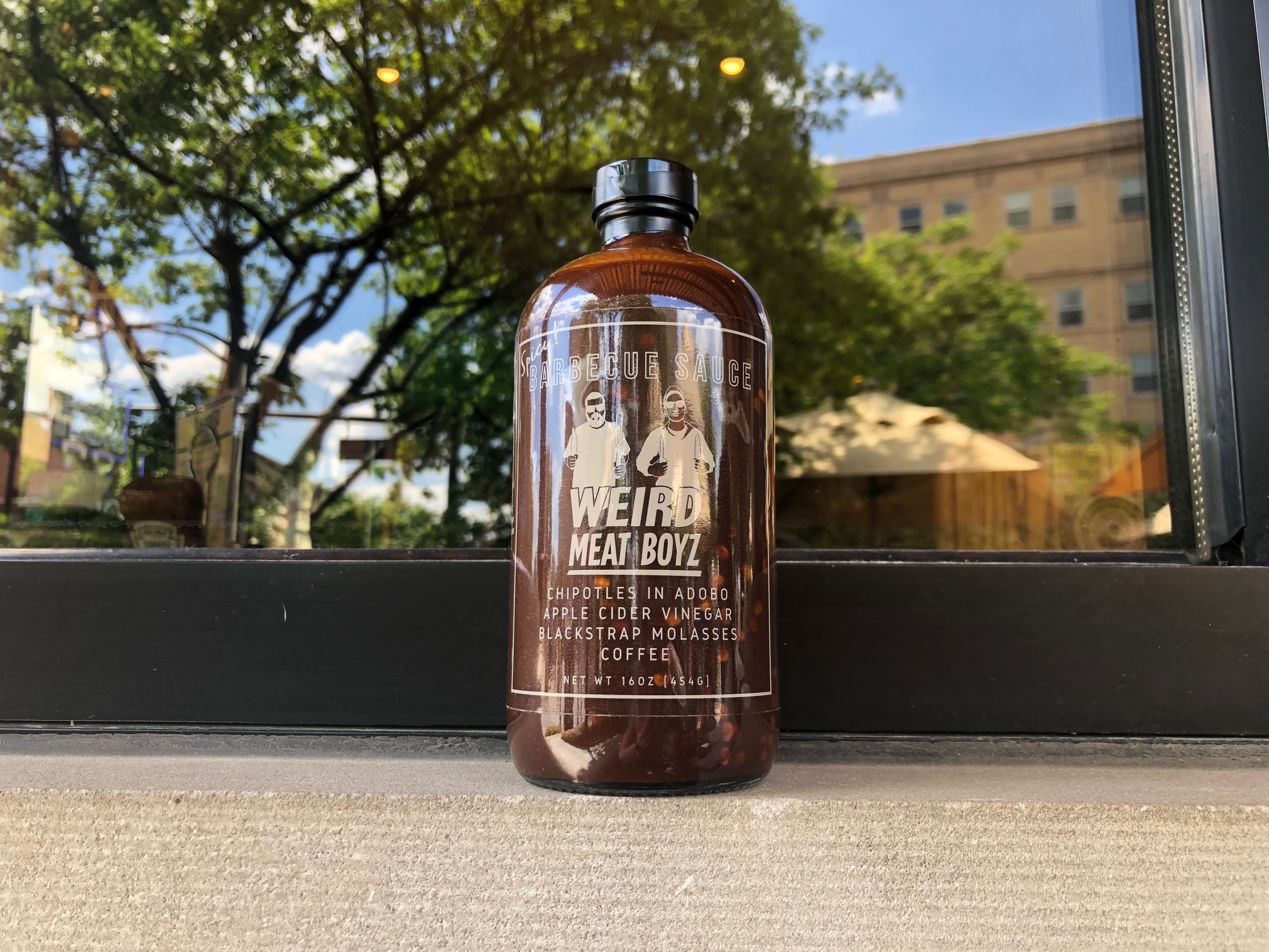 On a window ledge, a bottle of Weird Meat Boyz's BBQ sauce sits. In the window behind the bottle, there is a reflection of Downtown Champaign's trees and a bright blue sky. Photo by Alyssa Buckley.