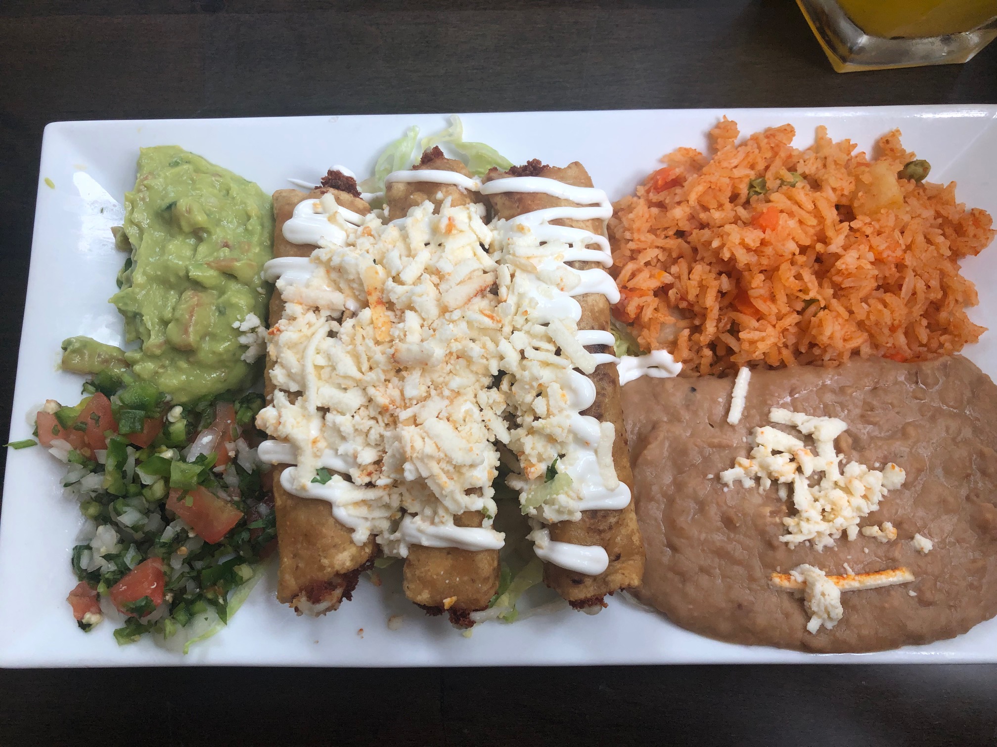 On a rectangular white plate, there is a pile of pico and guac beside three flautas covered in sour cream and cheese beside rice and beans. Photo by Alyssa Buckley.