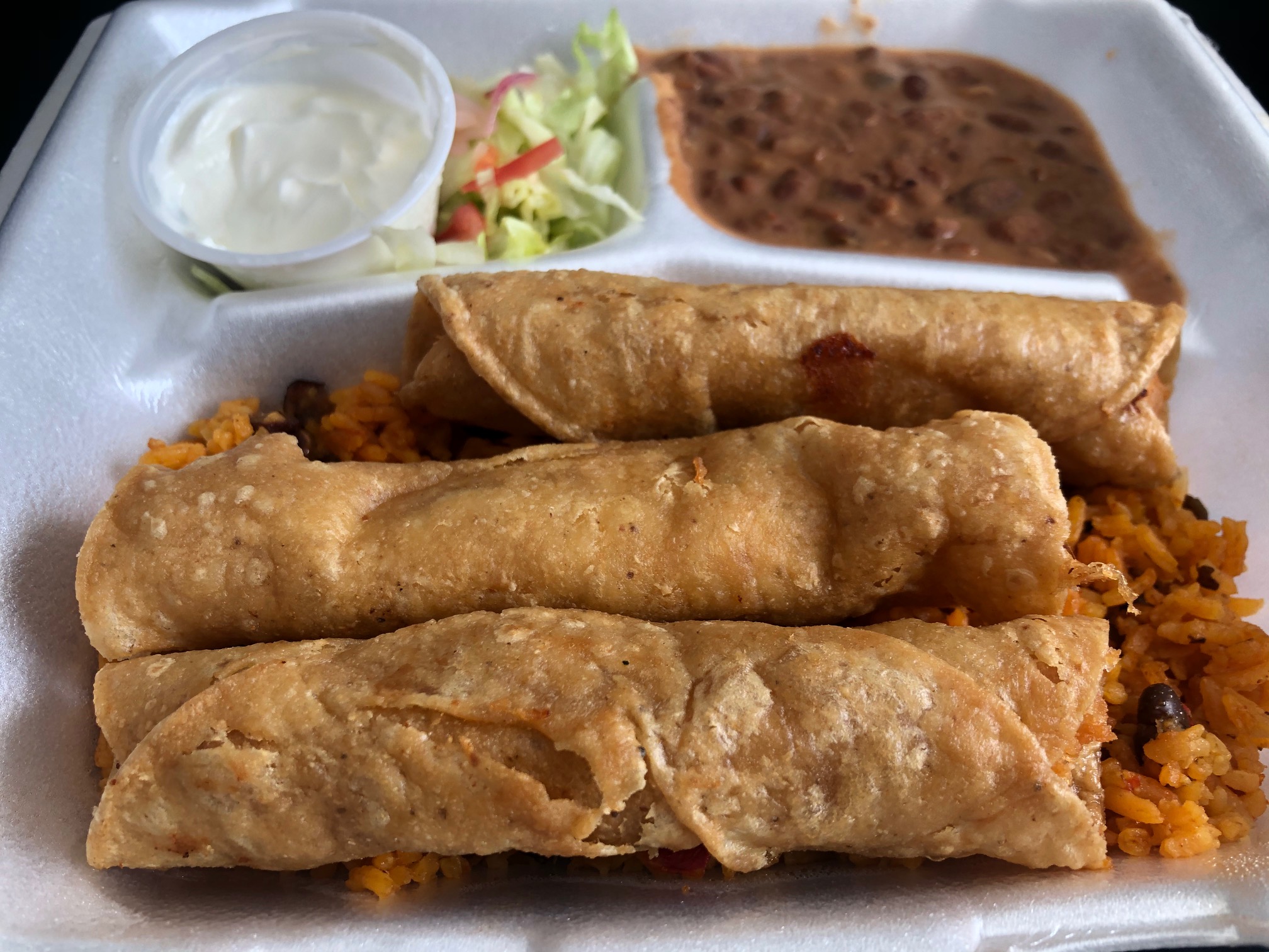 In a styrofoam container, there are three flautas atop rice with two dividers in the background with sour cream and lettuce on the left and beans on the right. Photo by Alyssa Buckley.