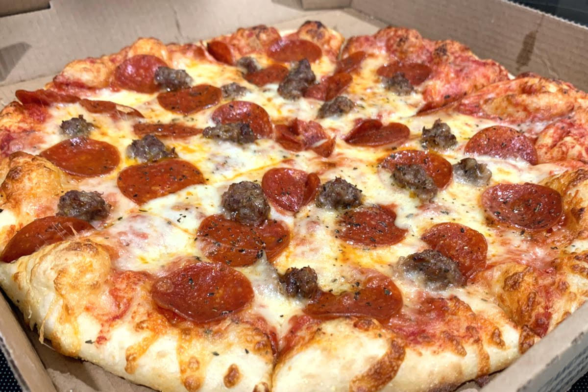 A photo of a medium-sized pizza in a carboard box. The pizza has a golden-brown crust with white cheese, red pepperoni and browned pieces of sausage. The pizza is speckled with black pepper and cut into slices. Photo by Megan Friend.