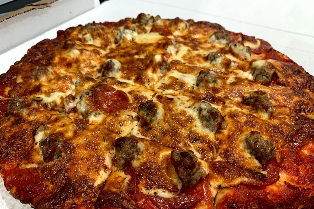 A photo of a medium-sized pizza in a cardboard box. The pizza has a thin crust with brown, crispy edges. There are large chunks of sausage and slices of pepperoni sticking out from the white, melted cheese. The pizza is cut into squares. Photo by Megan Friend.