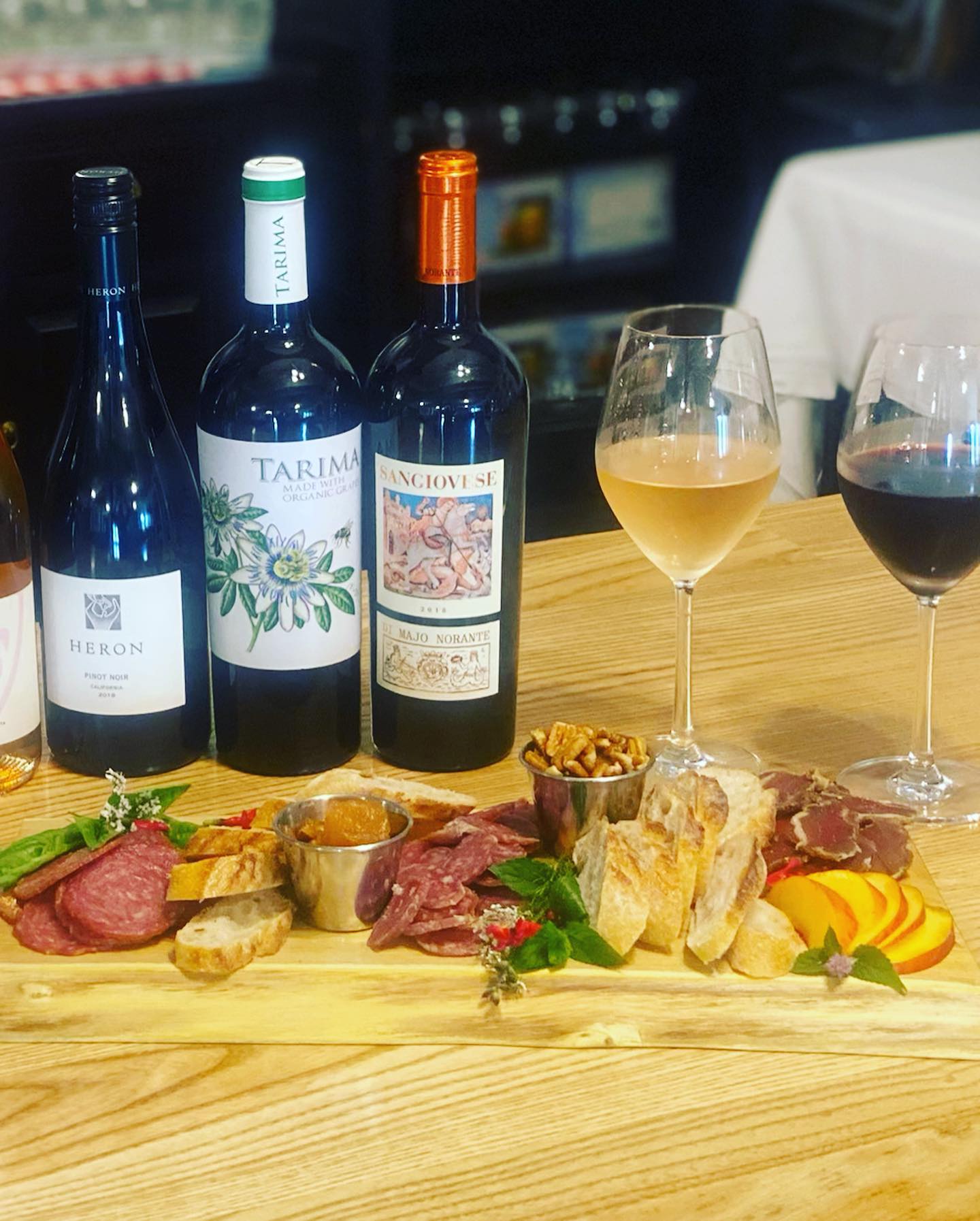 On a wooden table, there is a wooden board of charcuterie and three bottles of wine with two wine glasses behind it. Photo from Prairie Fruits Farm and Creamery's Facebook page.