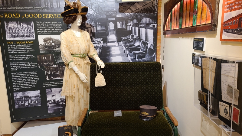 A museum display with a mannequin wearing a long beige dress and a large hat with feathers. It is standing next to a railway car seat, in front of a display about railroad history. Photo by Jaclyn Jones.