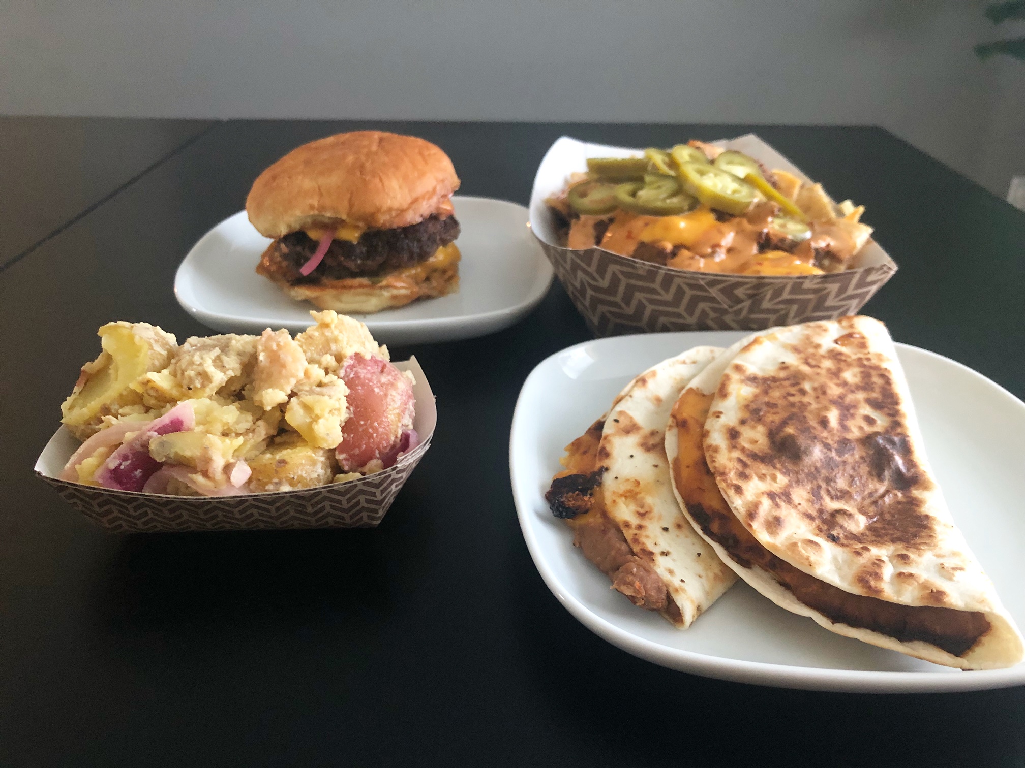 The author's dinner from the pop up food truck, starting with the bottom left: potato salad in a paper basket, above that a burger on a white square plate, beside that a paper basket of nachos, and below is a white square plate with two bean tacos on it. Photo by Alyssa Buckley.