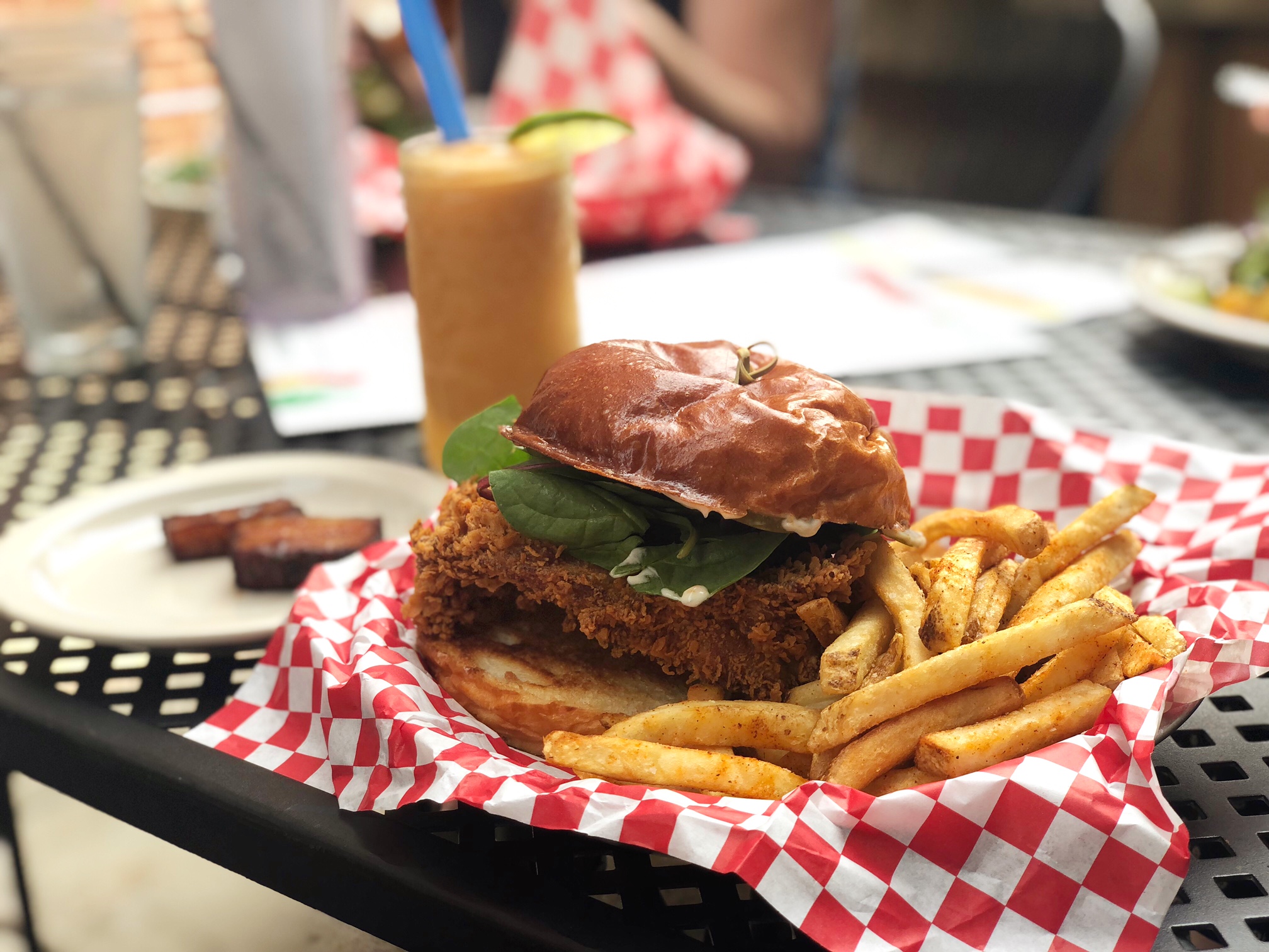 On an outdoor patio table, there is a plate with a chicken sandwich and skinny fries on a red-and-white checkered parchment paper. Beside the sandwich plate, there is a small white plate with two slices of pork belly. In the background, there is an orange slushie drink. Photo by Alyssa Buckley.