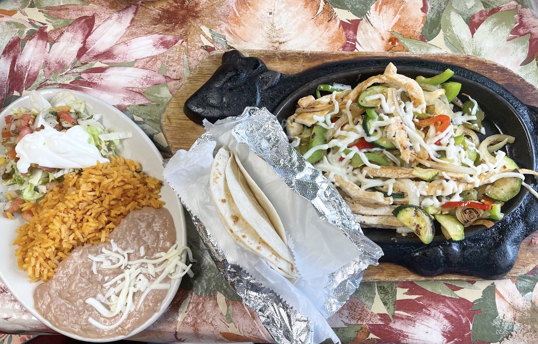 On a vibrant flowery tablecloth at Huaraches Moroleon in Urbana, there is a late of rice and beans with fajitas topping, a tin foil wrapped handful of flour tortillas, and chicken with veggies on a cow shaped cast iron plate. Photo by Alyssa Buckley.