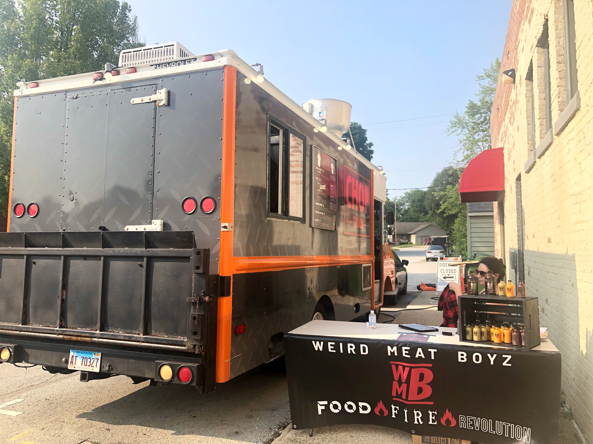A food truck facing away from the camera sits on the curb beside Champaign County Brewing Company. There is a table with a black tablecloth, and a white woman with brown hair working the table of hot sauces. Photo by Alyssa Buckley.