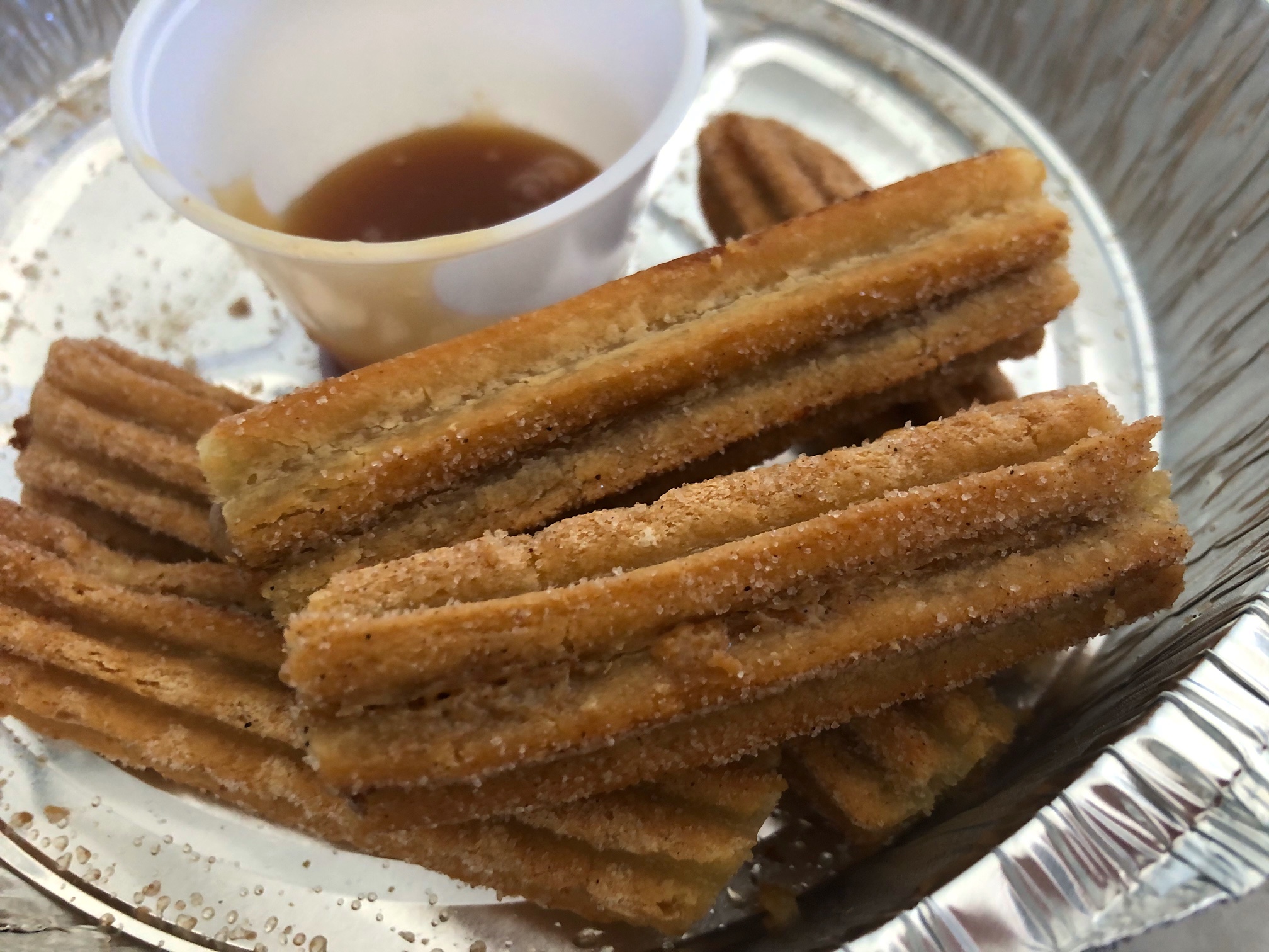 In a circular tin foil takeout container from El Toro II in Champaign, there are six miniature churros dusted in cinnamon sugar beside a small plastic cup with almost no caramel sauce in it. Photo by Alyssa Buckley.