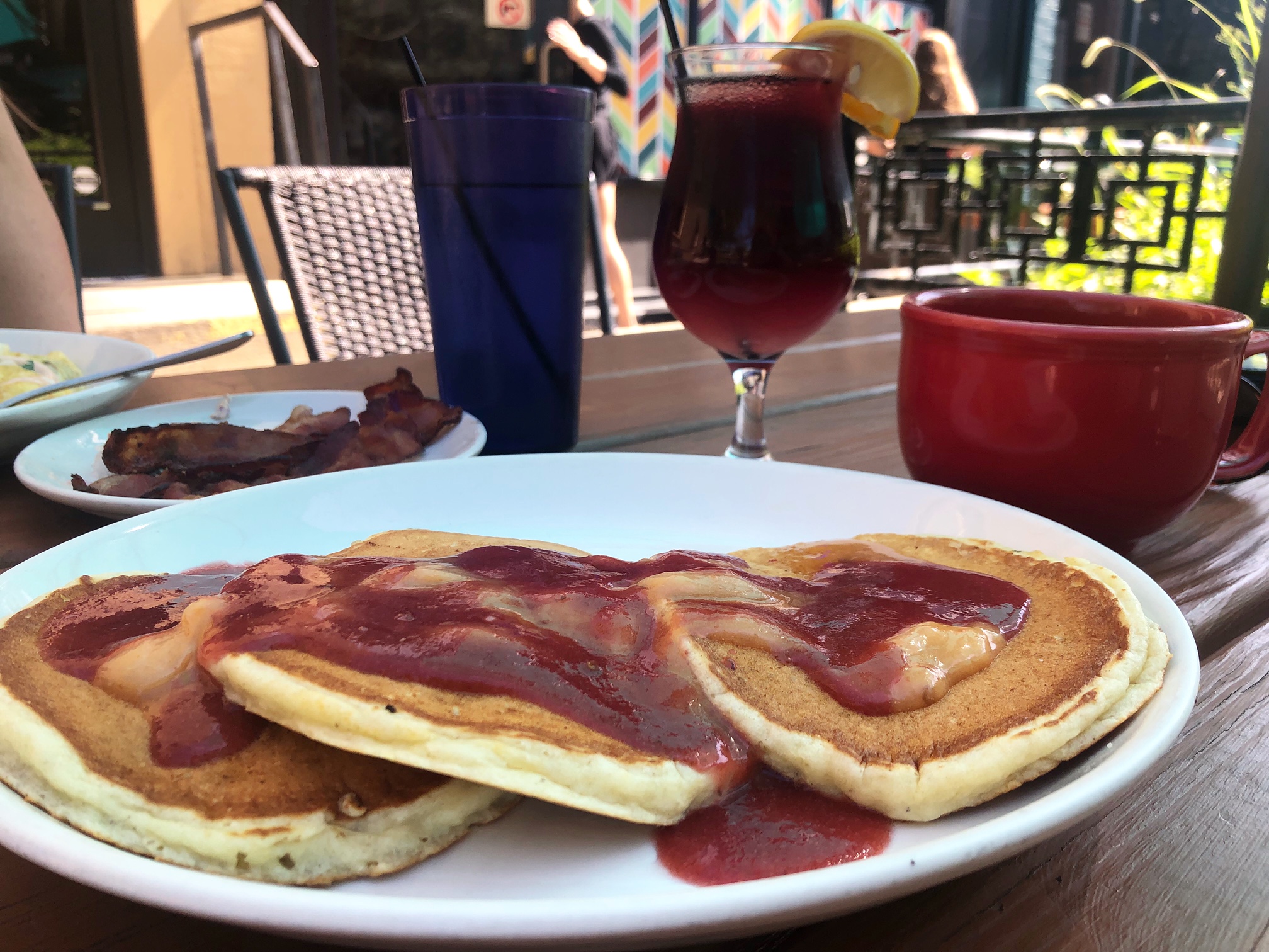 On a brown picnic table at the patio of Cowboy Monkey, there is a white plate with three pancakes on it. The pancakes have a dark pink syrup and a melting yellow butter. There is a hurricane glass of red sangria and a red mug of coffee behind the white plate of pancakes. Photo by Alyssa Buckley.