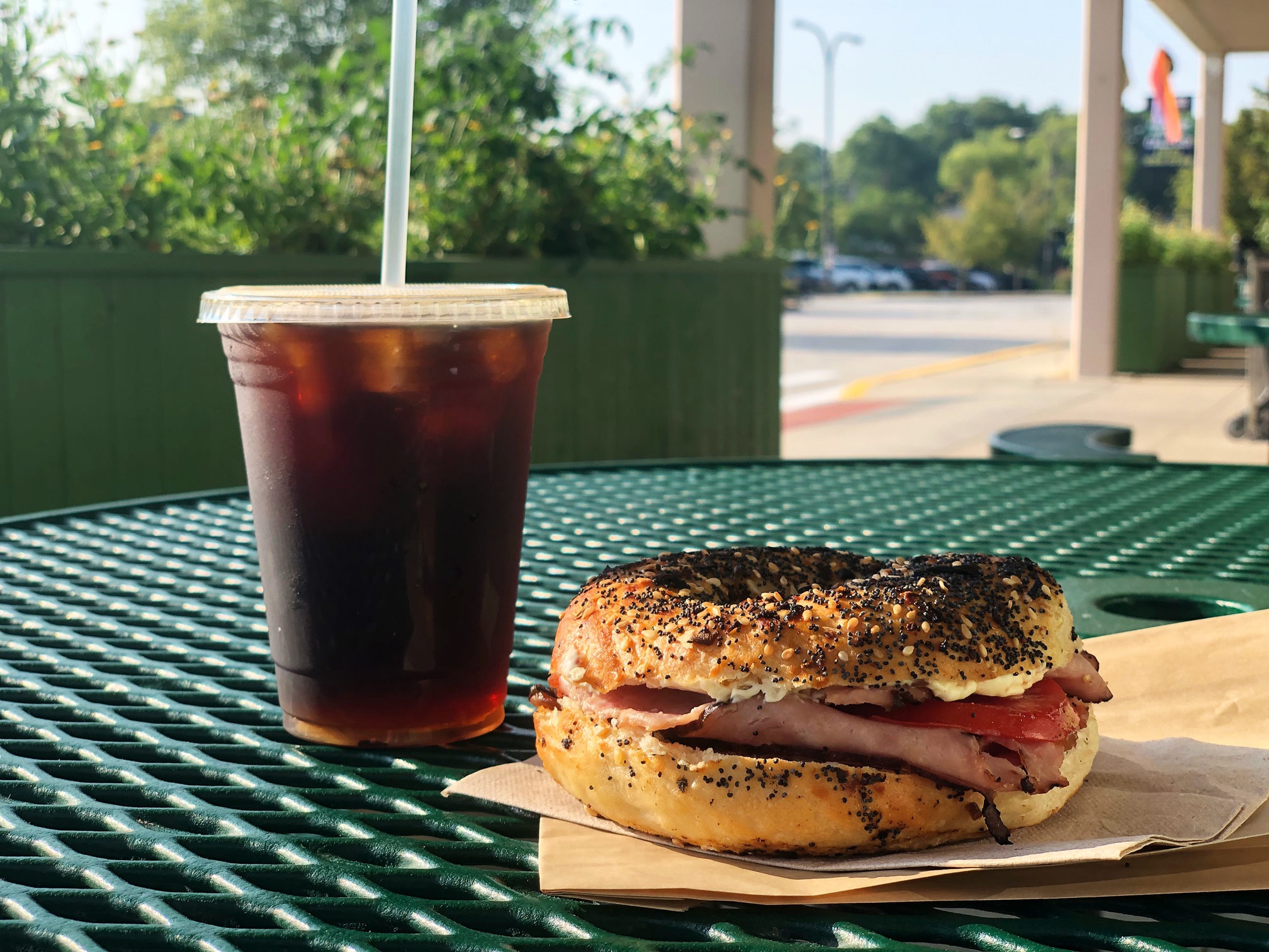 On a metal dark green table outside of the Common Ground Food Co-op in Urbana, there is a cold brew with a plastic straw in it beside a bagel sandwich on brown paper napkins. Photo by Alyssa Buckley.