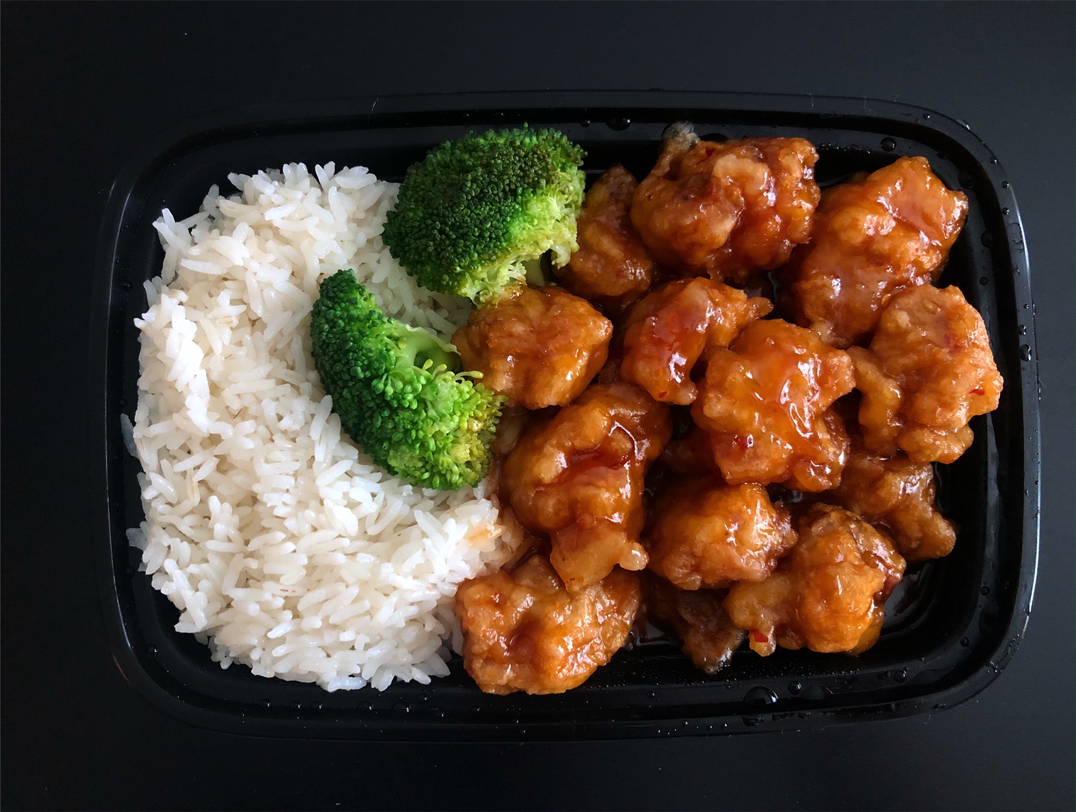 In a black plastic takeout container, there is white rice on the left side with broccoli bites beside and then a half container full of General Tso chicken. Photo by Alyssa Buckley.