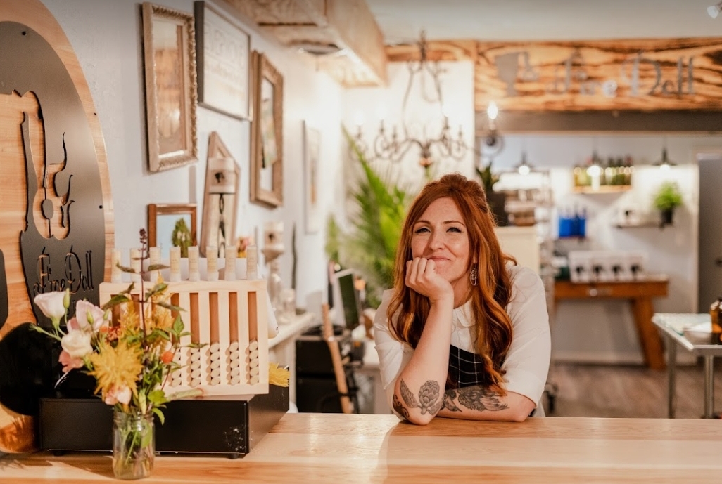 A woman with long reddish hair is leaning on a light wooden counter. There is a cash register and vase with flowers on the counter. Photo by Veronica Mullen.