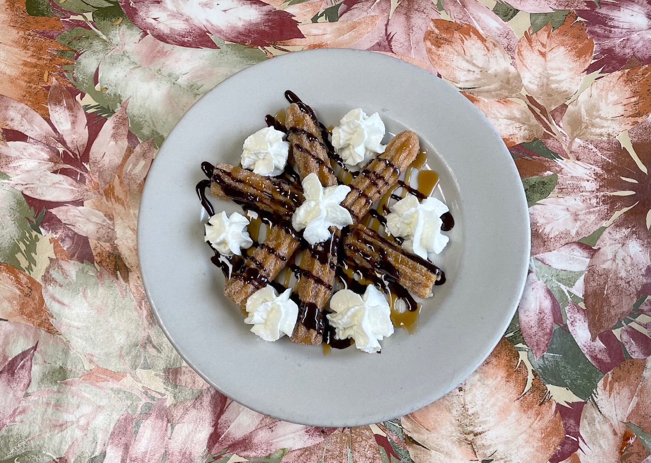 On a pale pink and green floral tablecloth at Huaraches Moroleon, there are six mini churros arranged in a star with puffs of whipped cream, chocolate drizzle, and a caramel drizzle. Photo by Alyssa Buckley.