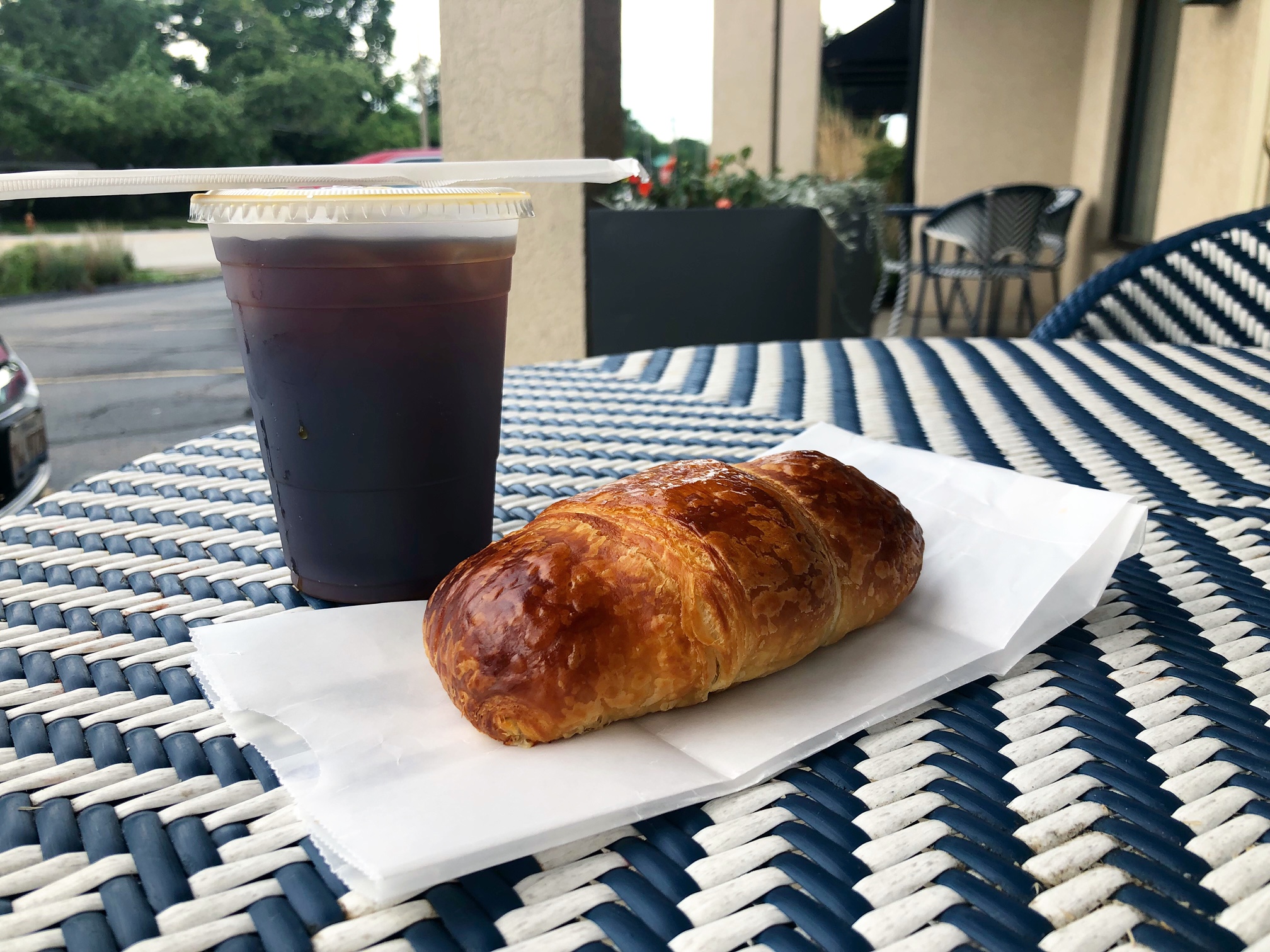 On the blue and white outdoor patio tables at Art Mart in Champaign, there is a plastic cup of dark cold brew with an unopened staw resting on top and a croissant on a white paper bag. Photo by Alyssa Buckley.