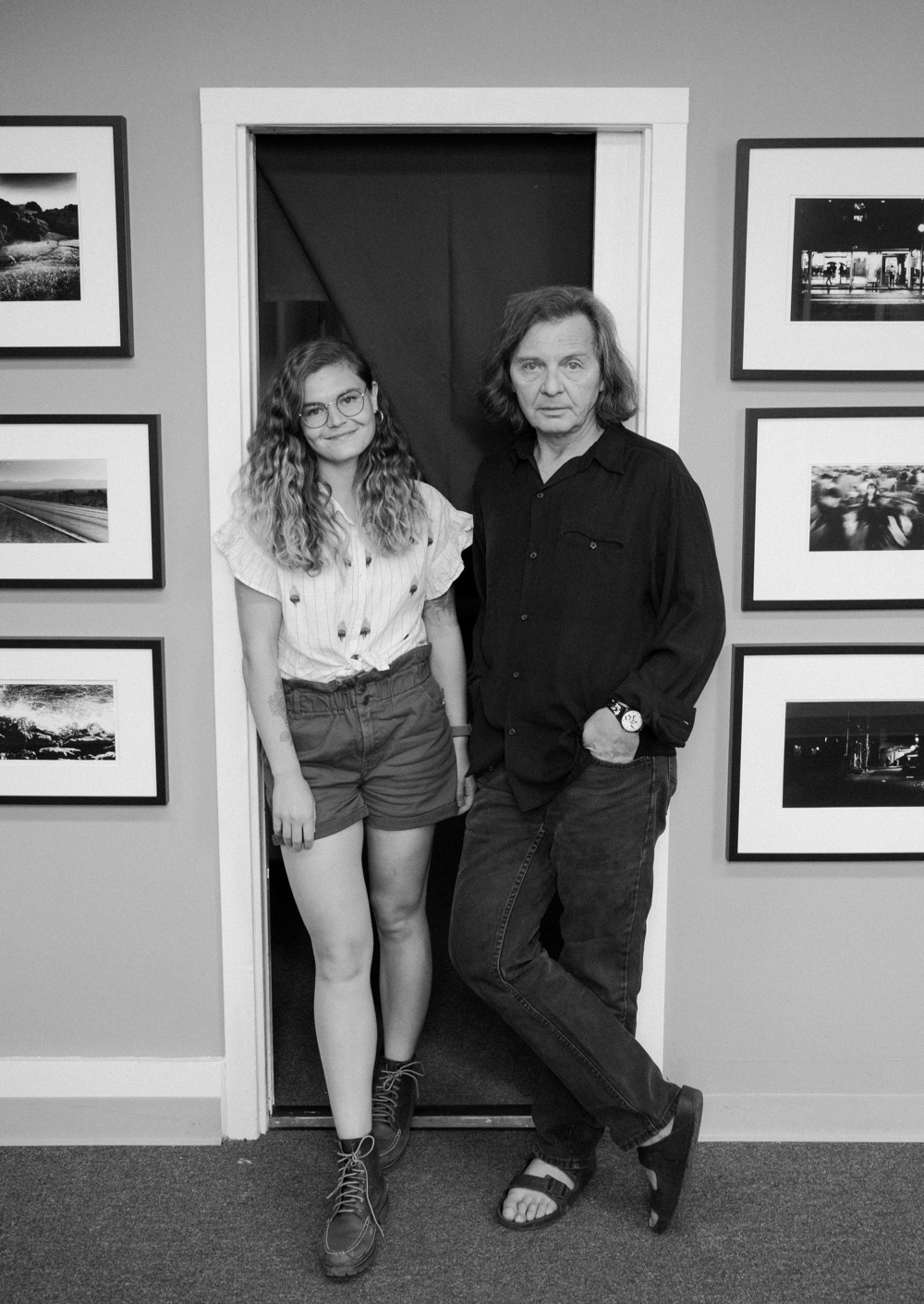 A black and white photo of Anna Longworth (left) and Lyosha Svinarski (right) standing looking at the camera in a door way