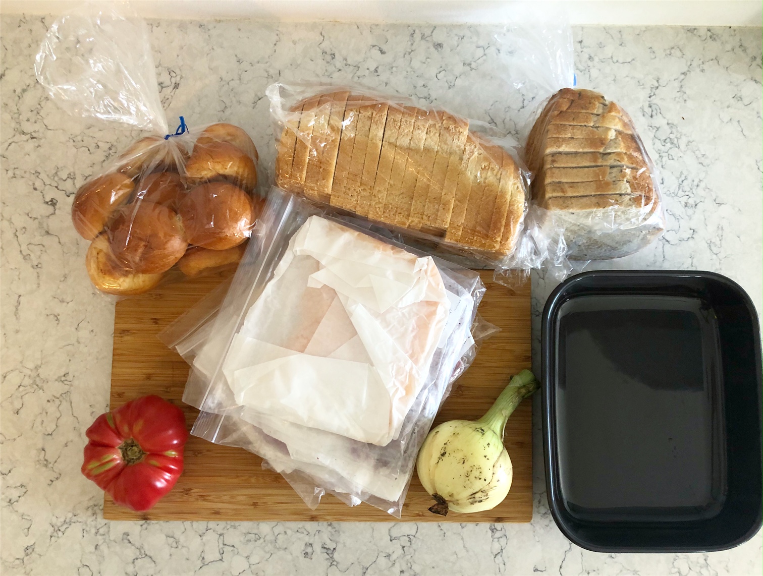 On a white veined counter, there is a wooden cutting board with plastic bags of bread, bags of sliced deli meat, a tomato, a dirty onion, and a black square ceramic container. Photo by Alyssa Buckley.