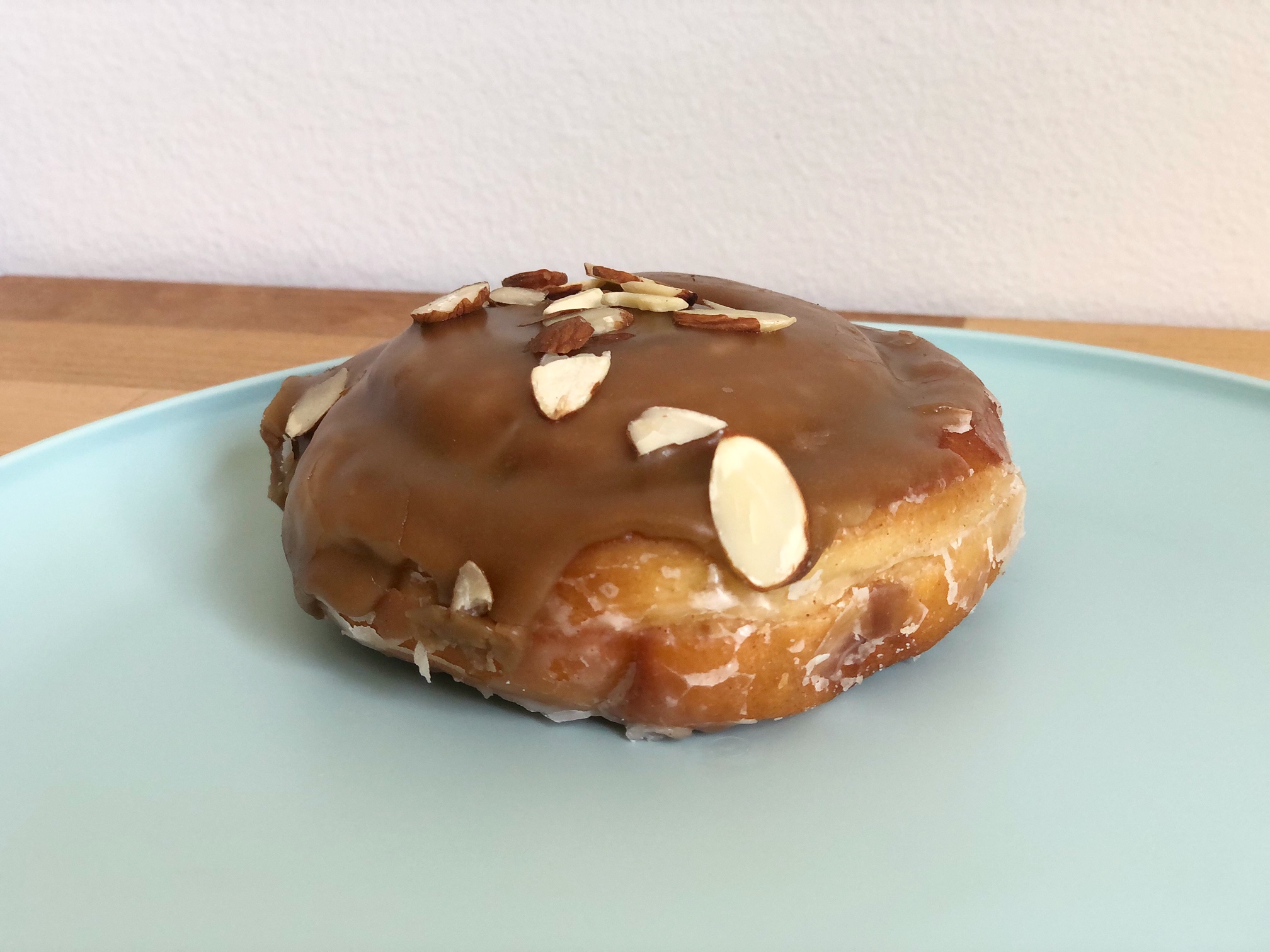 On a pale blue plate, there is a frosted donut with slivered almonds on top. The plate is on a wooden butcher block counter in front of a white wall. Photo by Alyssa Buckley.