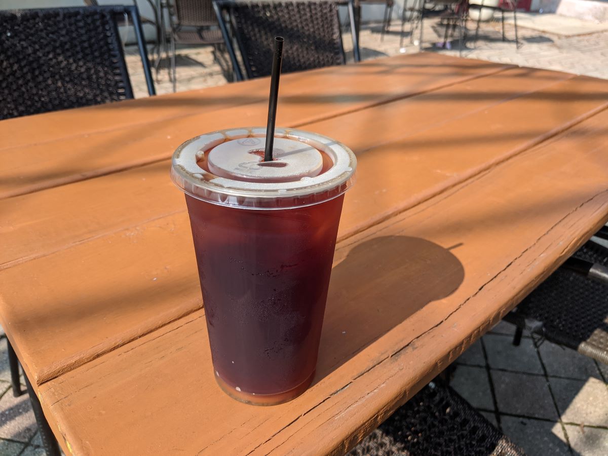 A reddish-brown tea in a clear plastic cup with a black straw. The cup sits on an orange-brown patio table in the sun. Photo by Tias Paul.