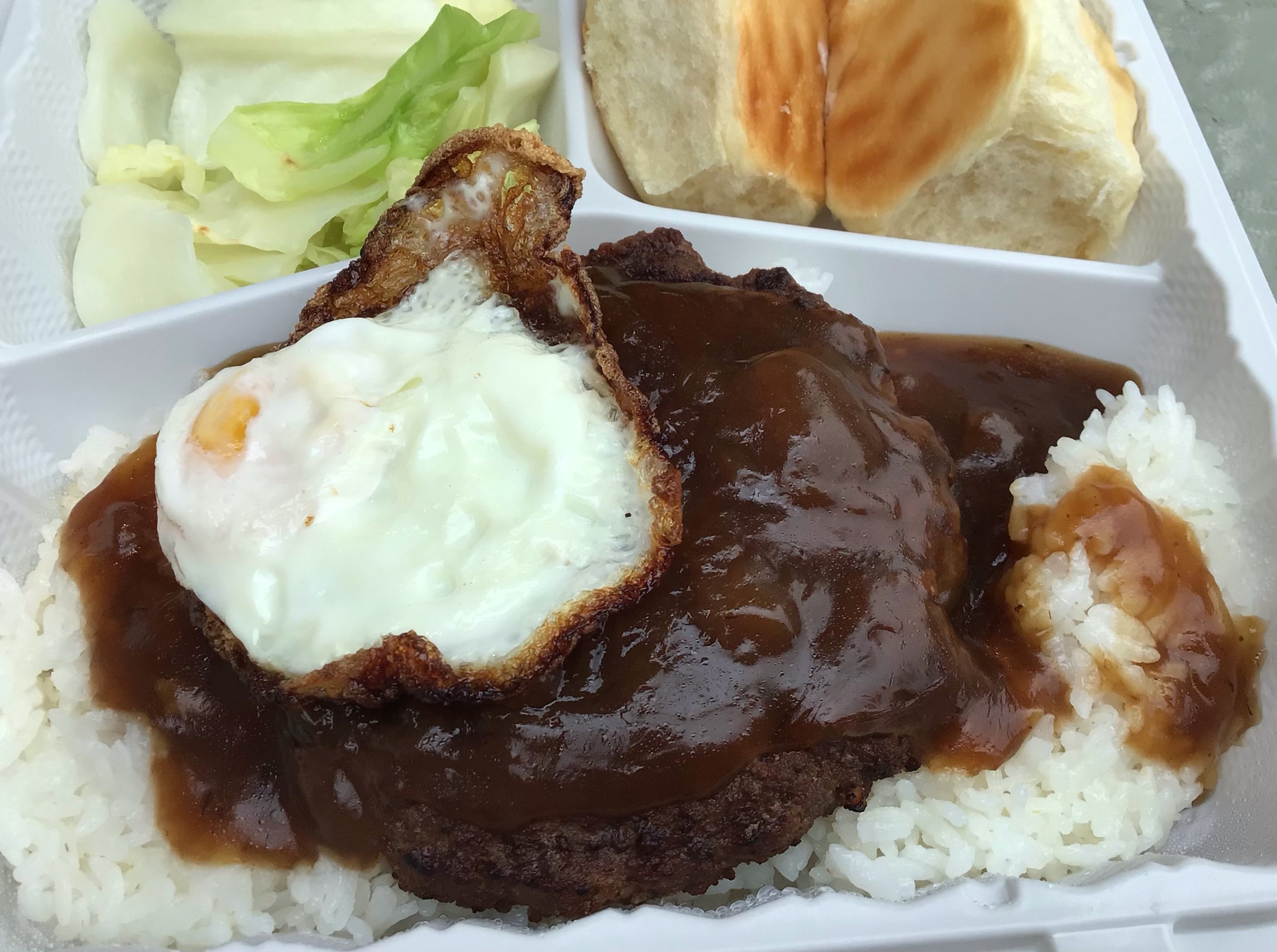 A white plastic to-go tray features a generous portion of loco moco (a seasoned hamburger patty covered in brown gravy and topped with a fried egg) atop a bed of steamed white rice in the main dish compartment. The side dish compartments contain, on the left, a single serving of cooked cabbage, and, on the right, two Hawaiian rolls.Photo by Rachael McMillan.