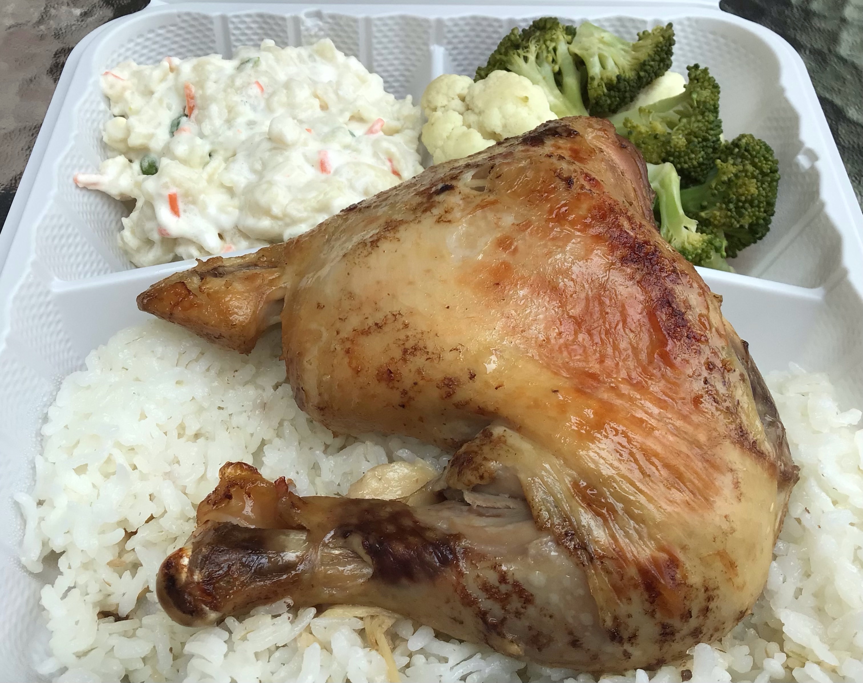 A white plastic to-go tray holds huli huli chicken (white rice topped with a roasted chicken leg quarter), a side of Hawaiian macaroni salad, and a side of steamed vegetables (broccoli and cauliflower are visible). Photo by Rachael McMillan.