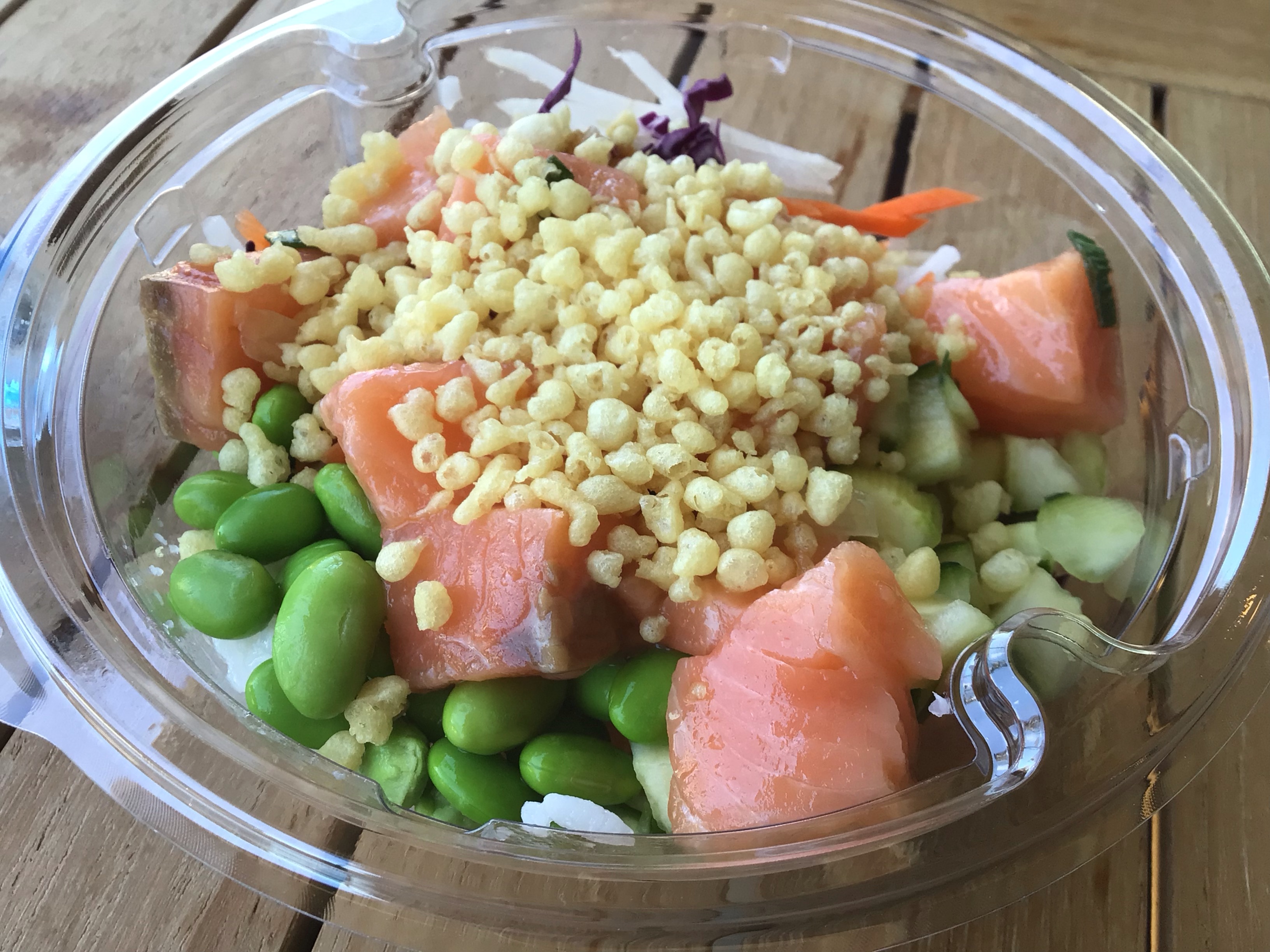 A clear plastic to-go container holds a salmon poke bowl: white sushi rice is topped with bright green edamame, shiny chunks of salmon poke, shredded cabbage and carrots, and small chunks of cucumber. The optional tempura crunch topping resembles rice cereal. Photo by Rachael McMillan.