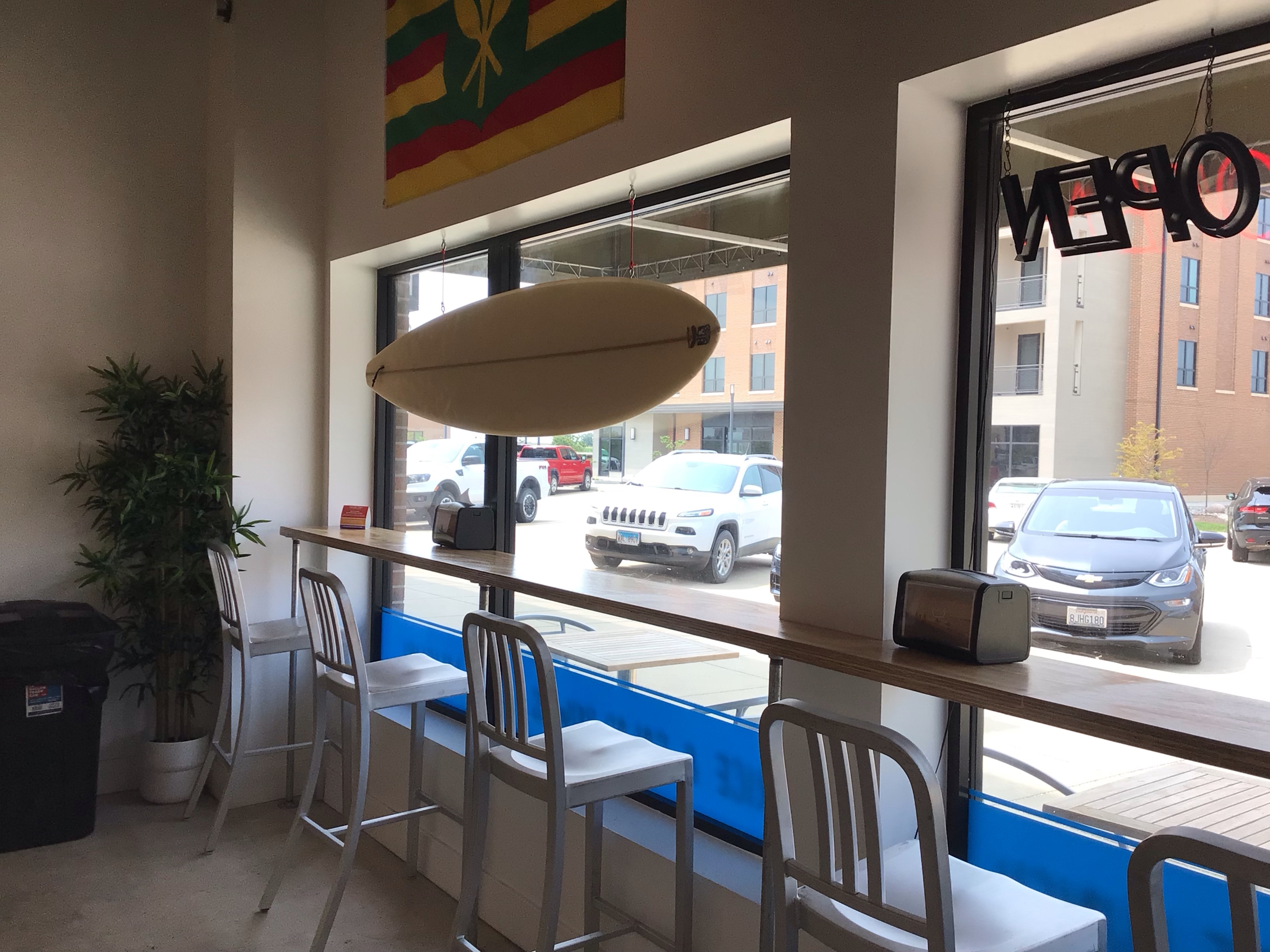 A view of Poke Shackâ€™s dine-in area shows a single, slim counter set up against the front windows of the restaurant. Four stools with metal frames/backs are pulled up to the counter, facing out into the street. Over one side of the counter hangs a cream-colored surfboard. The red, yellow, and green kanaka maoli Hawaiian flag hangs on the wall above the surfboard. Photo by Rachael McMillan.