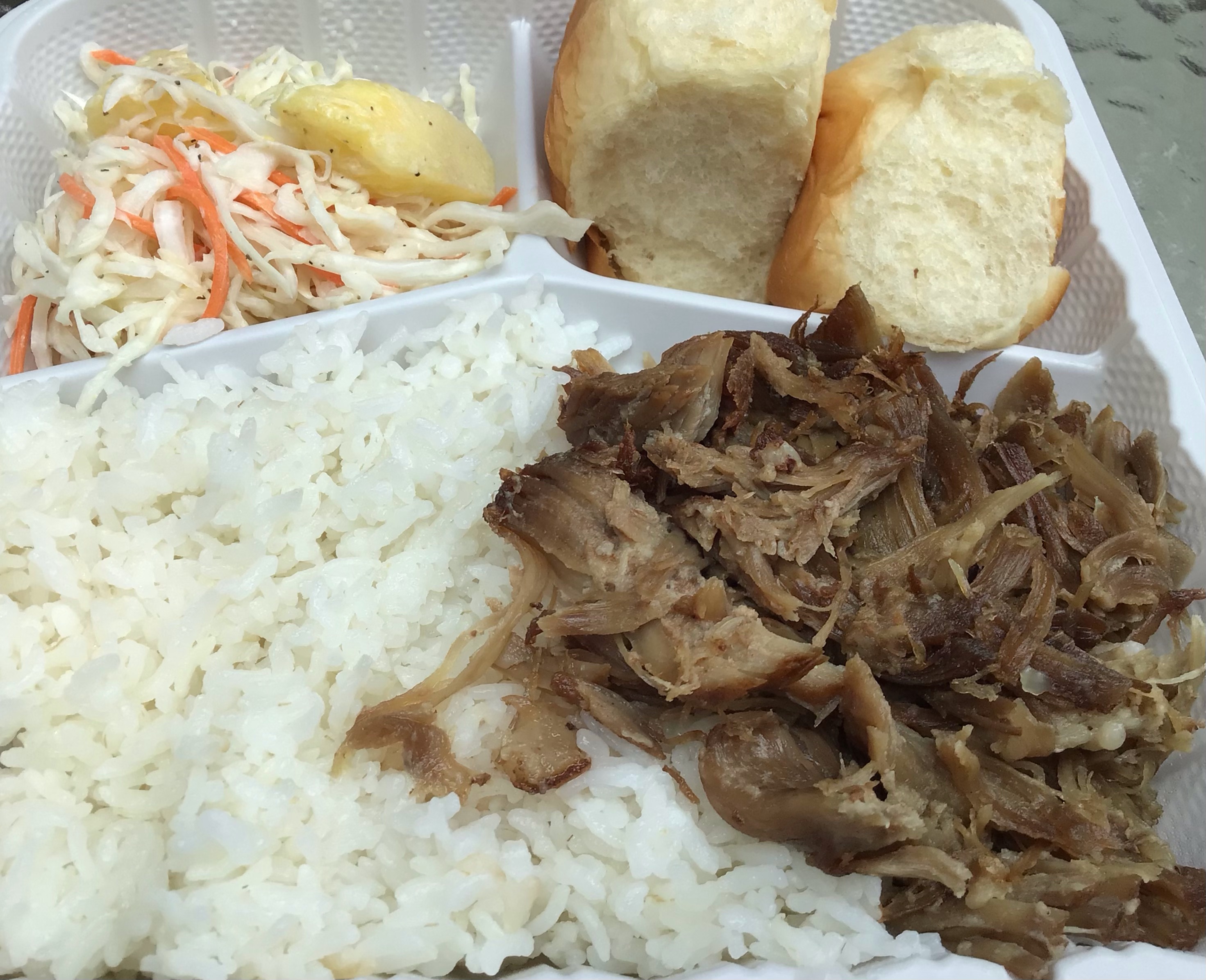 A white plastic to-go tray features a bed of steamed white rice in the main dish compartment, with a pile of slow-cooked, shredded pork set off to the right. The side dish compartments contain, on the left, a serving of pineapple coleslaw (shredded cabbage and carrots are visible, as is a large chunk of pineapple) and, on the right, two Hawaiian rolls. Photo by Rachael McMillan.
