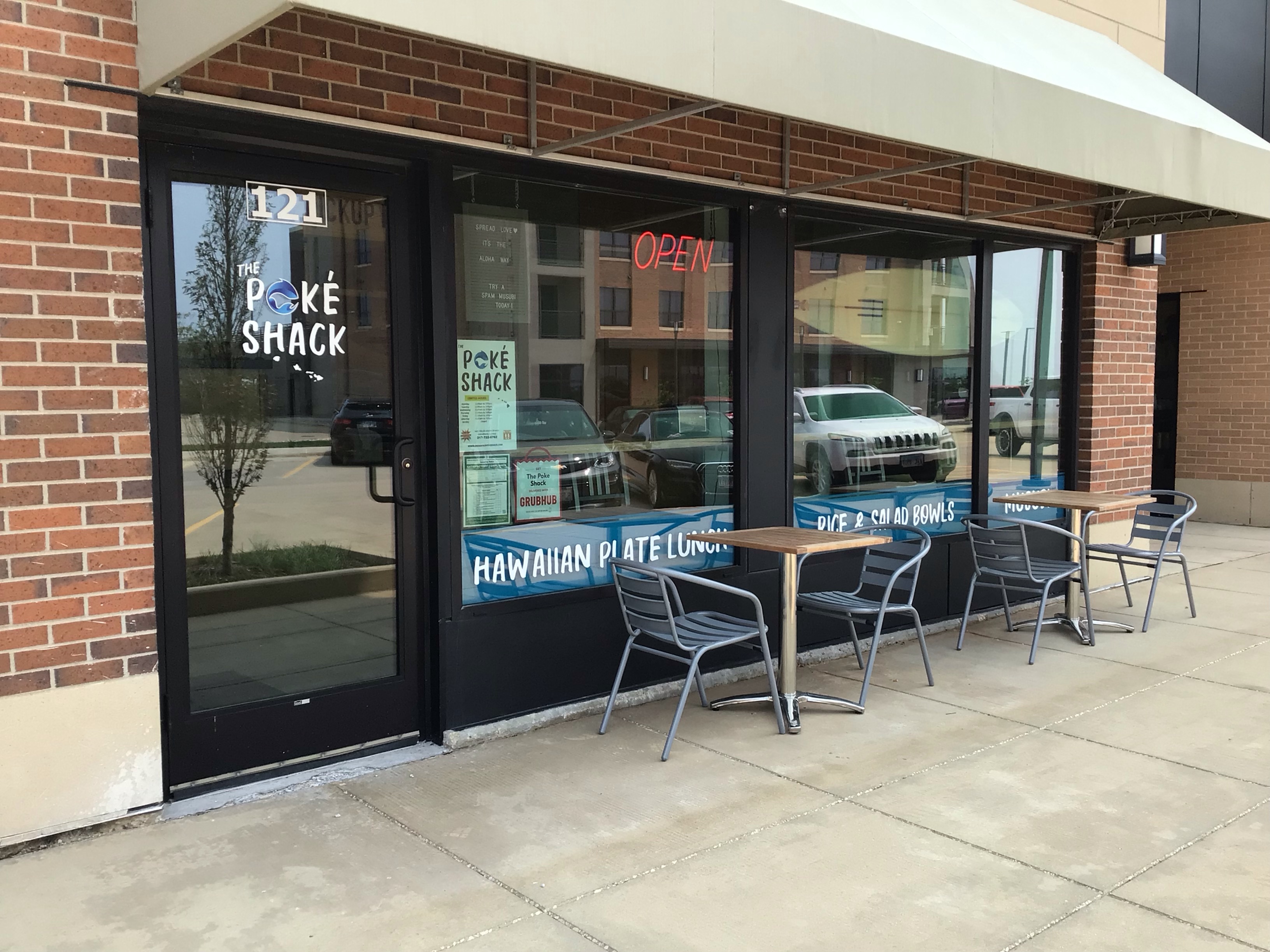 An exterior pic of the Poke Shack in Champaign at The Fields shows a small storefront with a door to the left of two large windows. In front of each window is a two-person outdoor dining set with metal chairs and wooden tables. â€œPoke Shack,â€ painted on the door, is the only signage. Photo by Rachael McMillan.