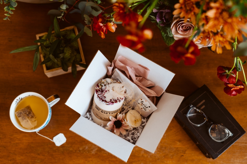 A square cardboard box is open, with large and small decorative candles, a flower, and tissue paper in it. A mug of tea sits next to the box, as well as a pair of glasses. Photo by Veronica Mullen.