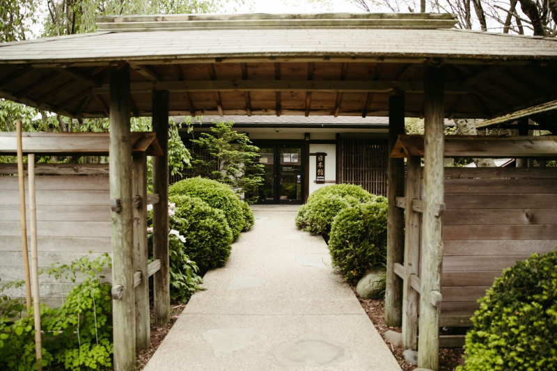 A view of the front entrance to Japan House. There is a sidewalk framed with manicured trees and bushes leading up to it. Photo by Anna Longworth.