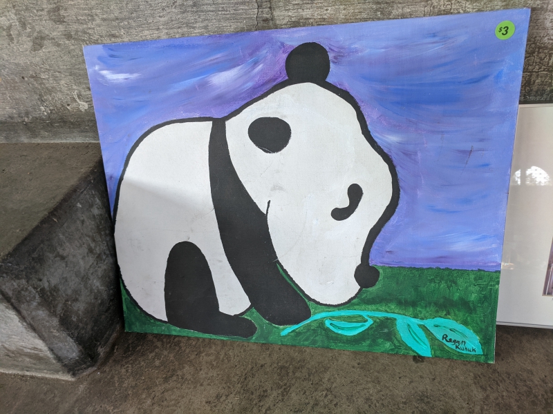 A canvas painted with a blue background and a black and white panda on a patch of grass with a stem of green bamboo. . Photo by Tom Ackerman.
