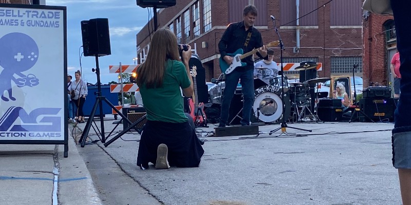 A man is playing a guitar, surrounded by other band members and equipment on a street. There is a woman kneeling in the foreground taking a photo. Photo by Julie McClure.