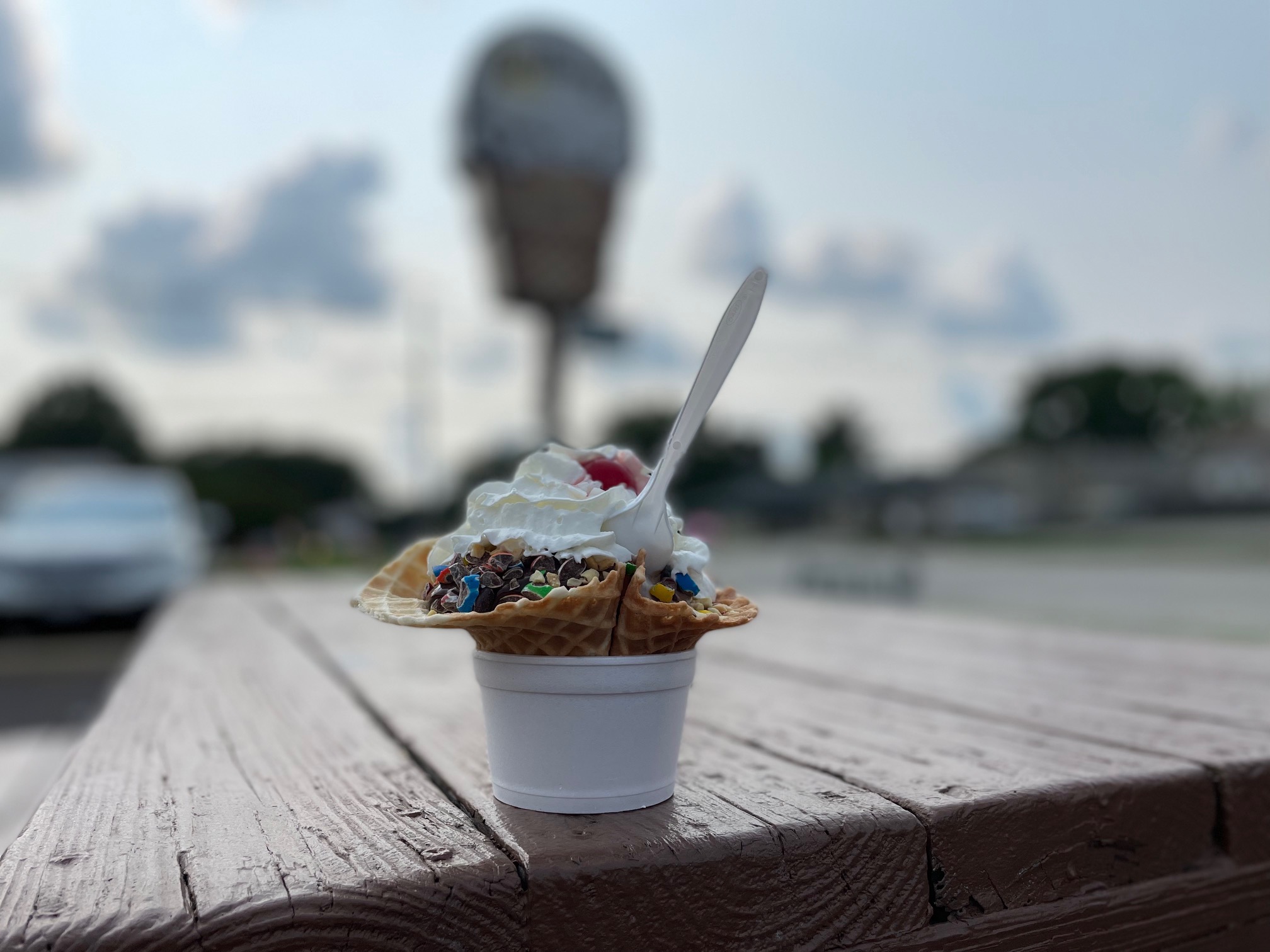 On a brown picnic table outside Jarlings, there is a white styrofoam cup with a waffle cone inside with ice cream, hot fudge, whipped cream, and a cherry on top with a white plastic spoon in it. Photo by Alyssa Buckley.