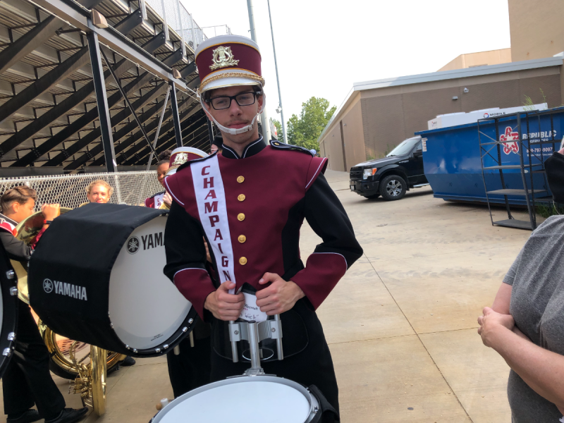 Central High School junior boy in a maroon and black marching band uniform is holding a snare drum. He is wearing thick-brimmed glasses. He is standing near bleachers and a chain-linked fence. Photo by Joanna Wozniak