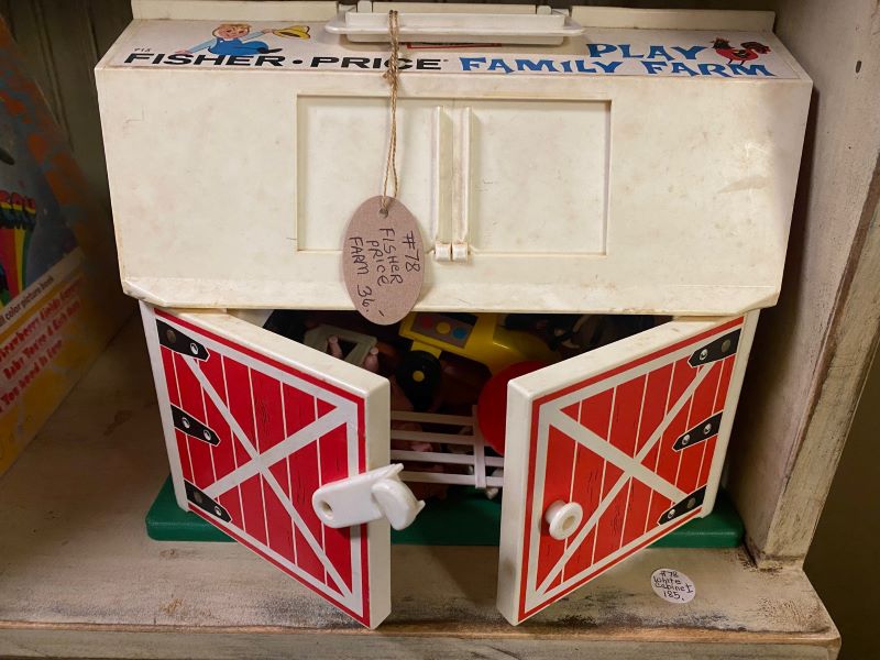 A toy barn with a white plastic roof with a red base. The barn doors are slightly open and there are plastic farm toys inside. It's sitting on a brown wooden shelf. Photo by Julie McClure.