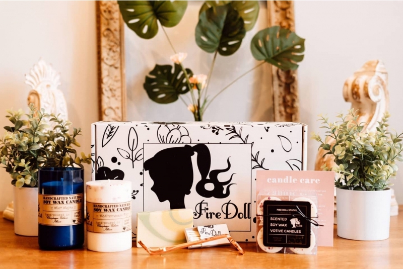 A rectangular box with black and white designs and Fire Doll Studios on the label. It is surrounded by different sizes and colors of candles, and a bar of soap. Photo by Veronica Mullen.