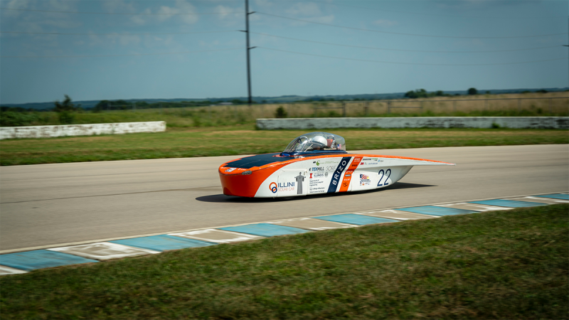 Solar-powered Brizo races through the scene as the background blurs from the speed. The car is white with blue and orange accents, its lower half is covered in advertisments and acknowledgements for sponsorships. Inside sits a student wearing a white helmet, visible through the clear dome of the cockpit in the center of the car. Photo provided by Illini Solar Car. 