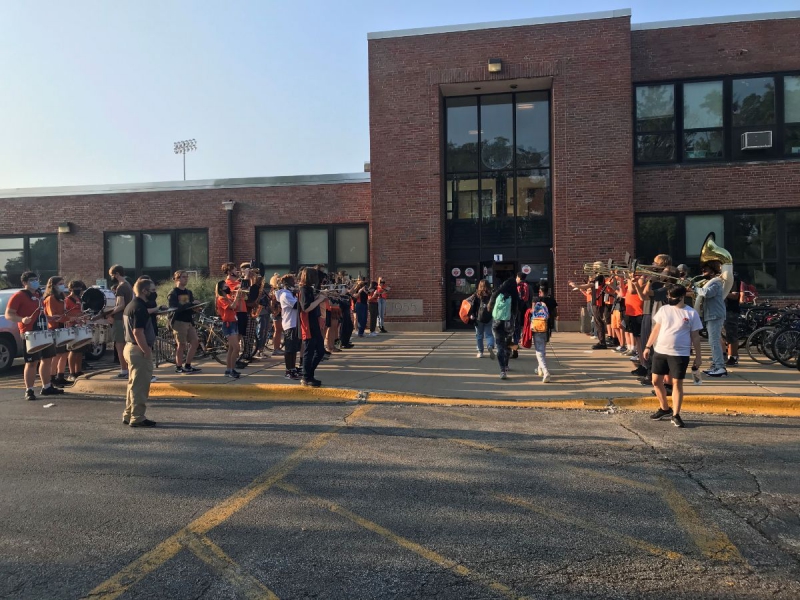 Urbana High School marching band members dressed in casual black and orange clothes are playing music as students walk into the building on the first day of school. Building is brown and red brick, and the sky is clear and sunny. Photo by Stacey Peterik.