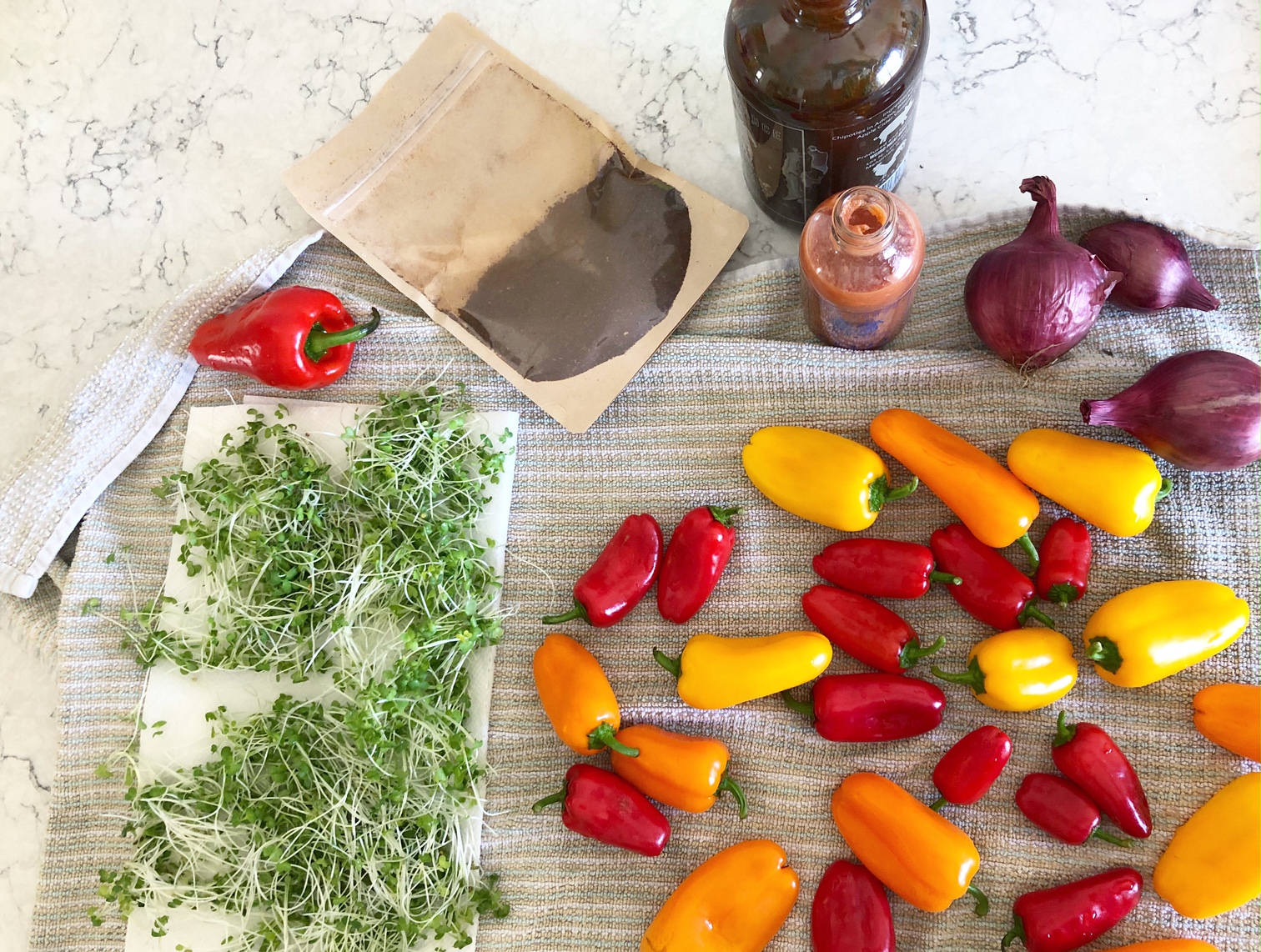 An above shot of the author's market purchases: microgreens on a paper towel, a bright red paprika pepper, a package of paprika seasoning, several small red onions, and a lot of red, orange, and yellow little peppers. Photo by Alyssa Buckley.