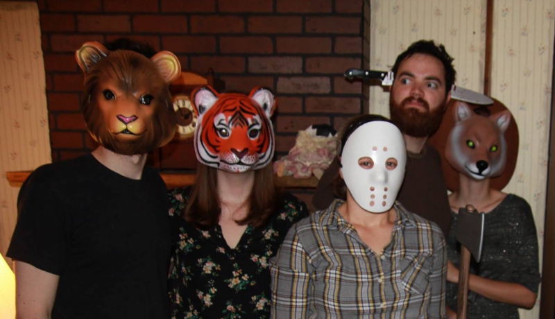 Five people are posing for a photo in front of a fake brick wall. Three are wearing animal masks, one is wearing a ski mask, and the writer has a fake knife through the head. Photo by CU Adventures in Time and Space.