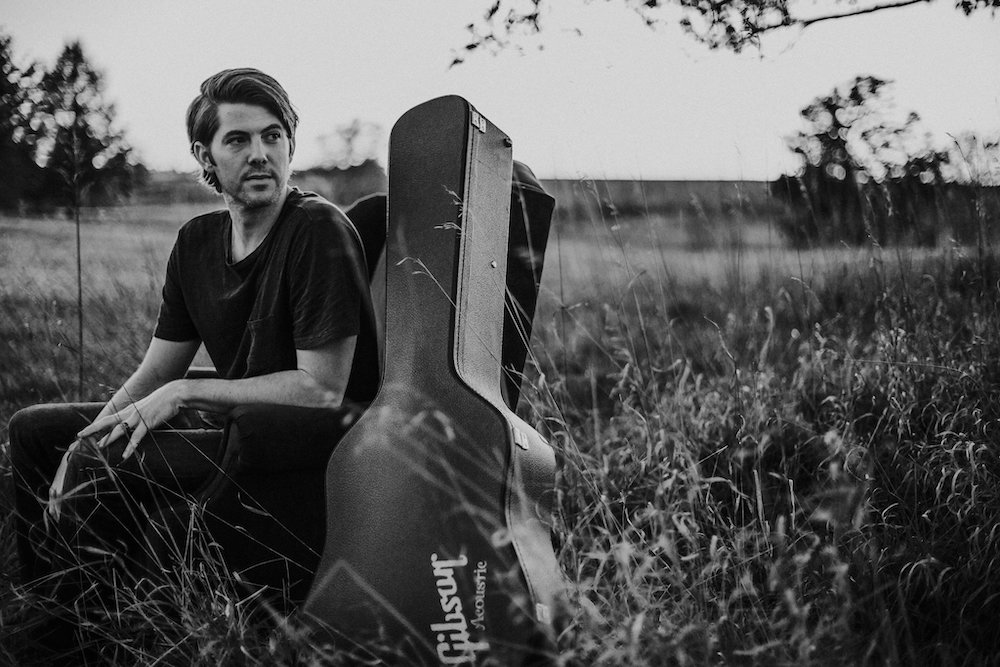 Black and white portrait of Dan Hubbard next to his guitar case as he looks into the distance. He is sitting in a prairie. 