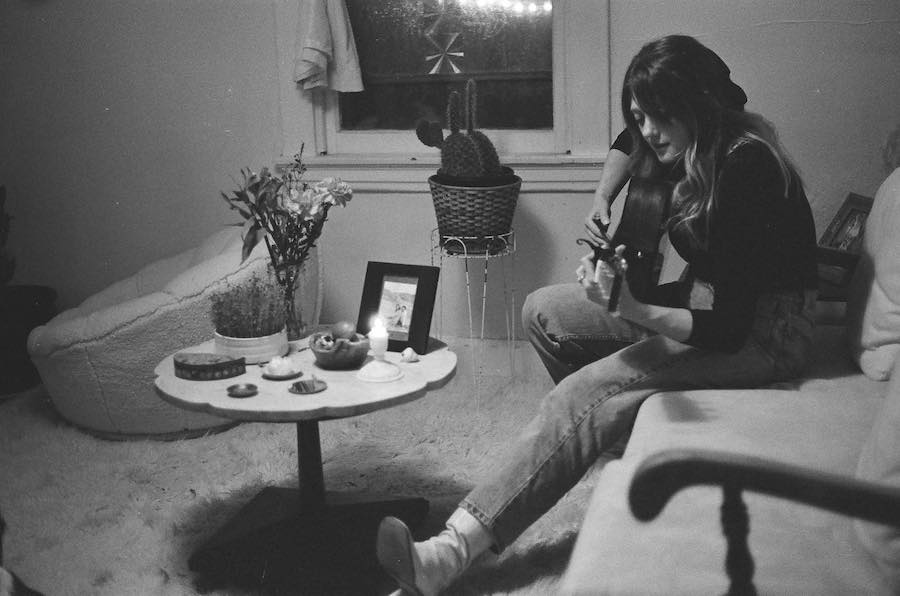 Image of musician Cara Louise sitting on a couch playing guitar. The table in front of her has a photo and candle. 