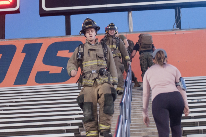 Two men in full firefighting gear walk down the steps of memorial stadium followed by a group of people in combat fatigues and carrying backpacks full of weights. Photo by Justin Malone.