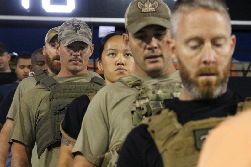 A woman in combat fatigues looks up surrounded by men in combat fatigues, Kevlar vests, and donning hats as they all walk down the stairs of memorial stadium. Photo by Justin Malone.
